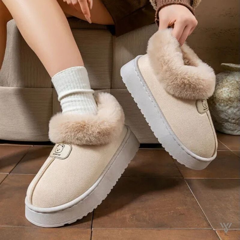 Beige snow boots slippers with fluffy fur trim worn indoors on a tiled floor.
